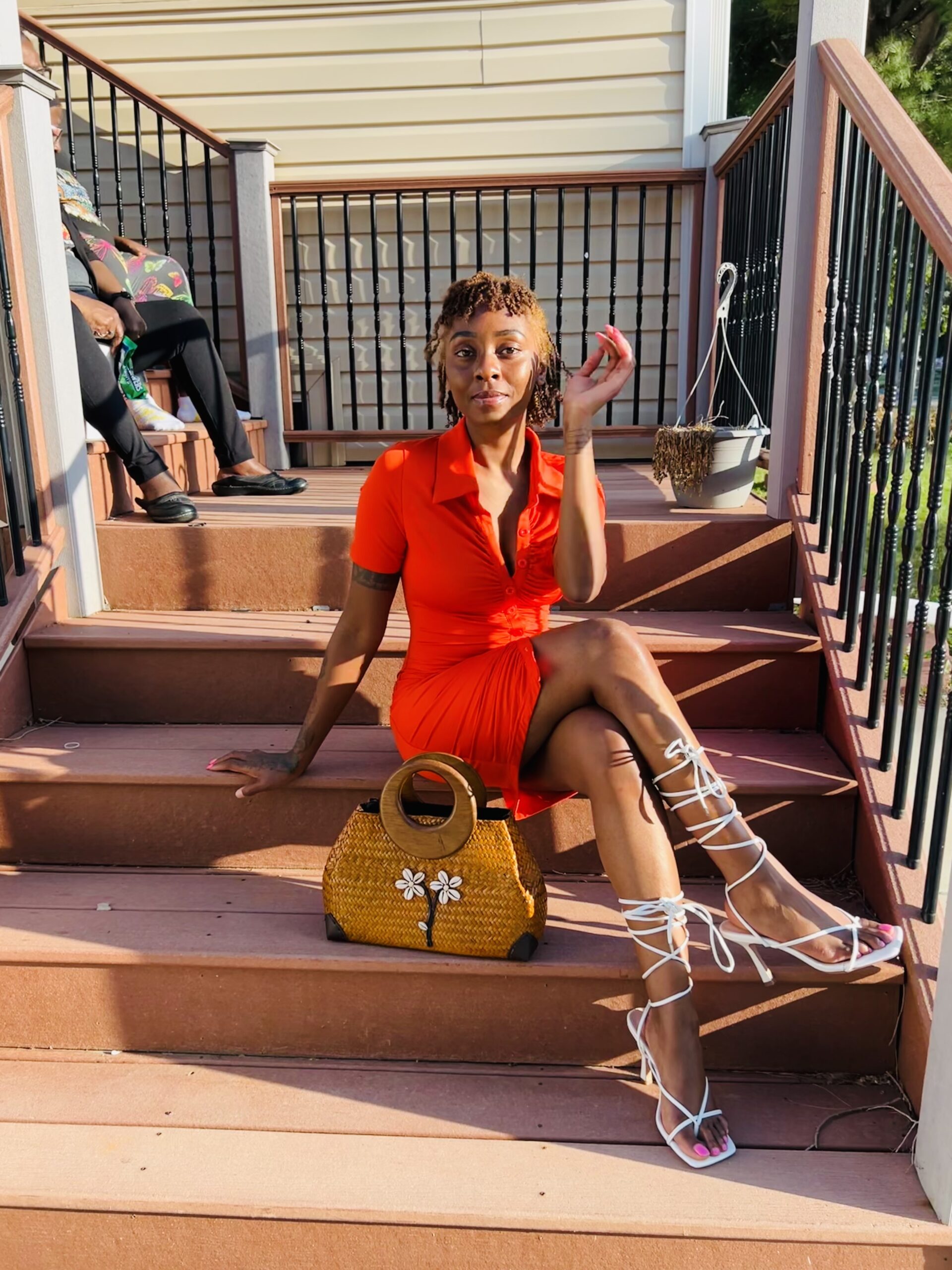 Content creator Elitrise seated on porch steps in a red-orange midi dress and white lace-up sandals with natural curls